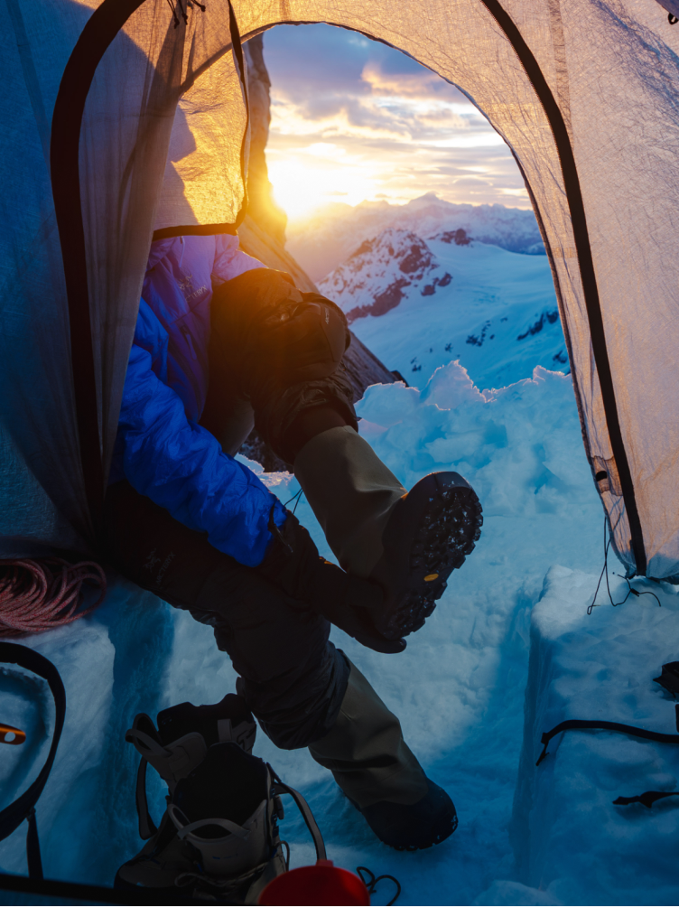 From inside a tent, a person's legs in winter boots face a bright sunrise illuminating a snow-covered mountain range.