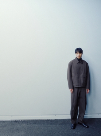 A man in a dark brown jacket and trousers with a black turtleneck, standing against a light wall.