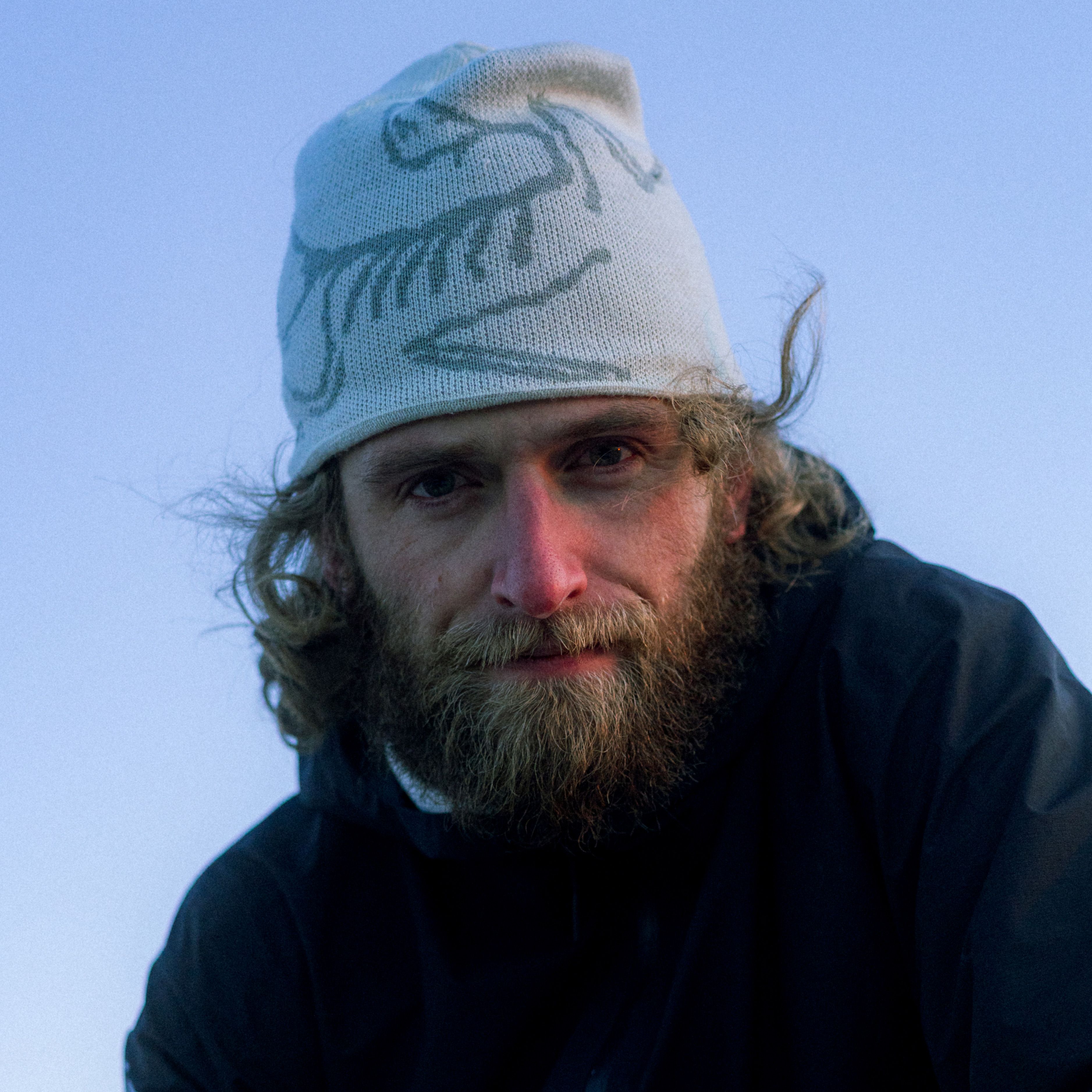 A man with a full beard and curly hair wearing an Arc'teryx beanie looks directly at the camera against a blue sky.