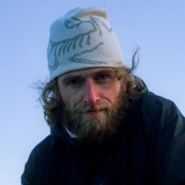 A man with a full beard and curly hair wearing an Arc'teryx beanie looks directly at the camera against a blue sky.