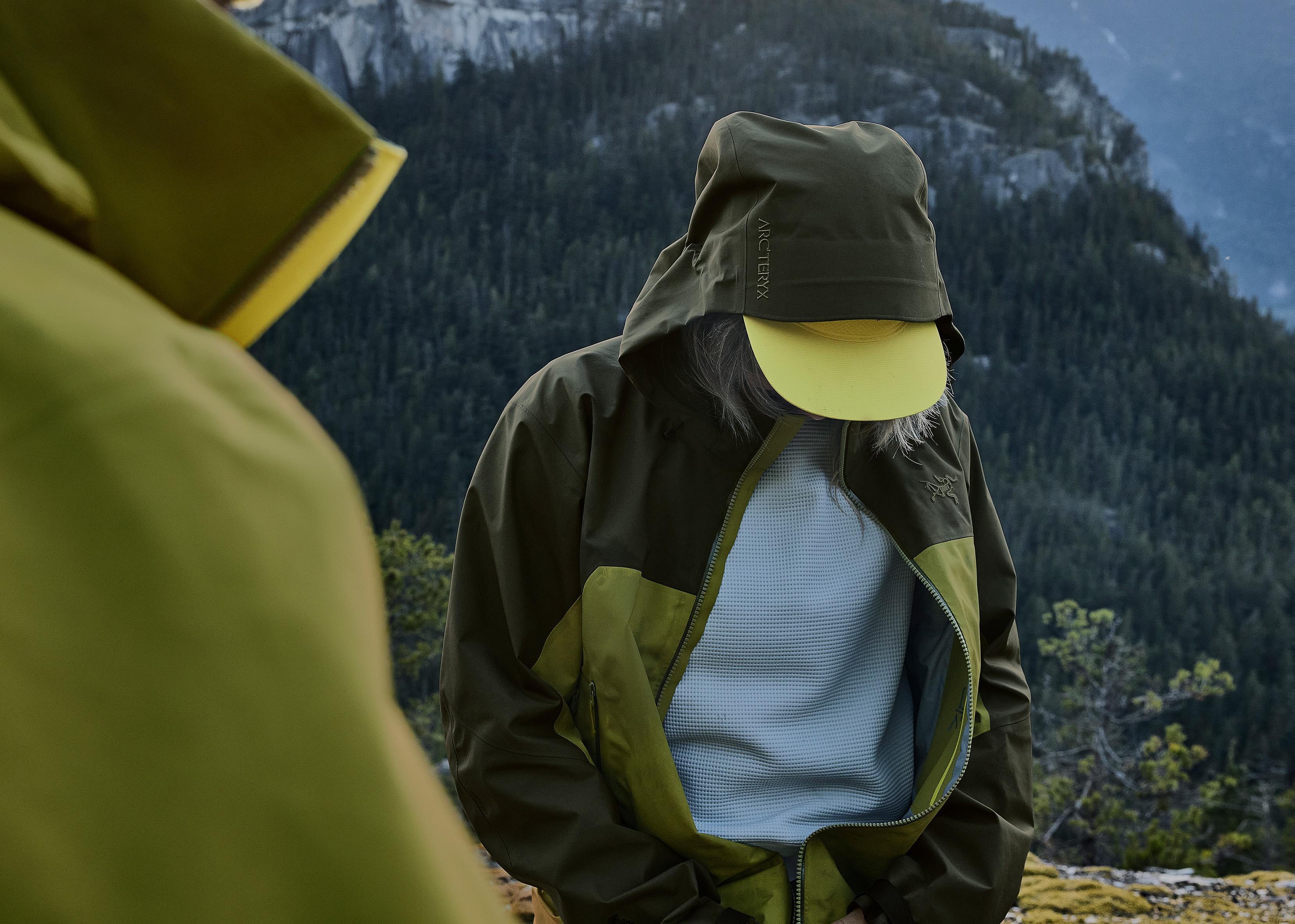 a person wearing a green jacket and a yellow hat is standing in front of a mountain .
