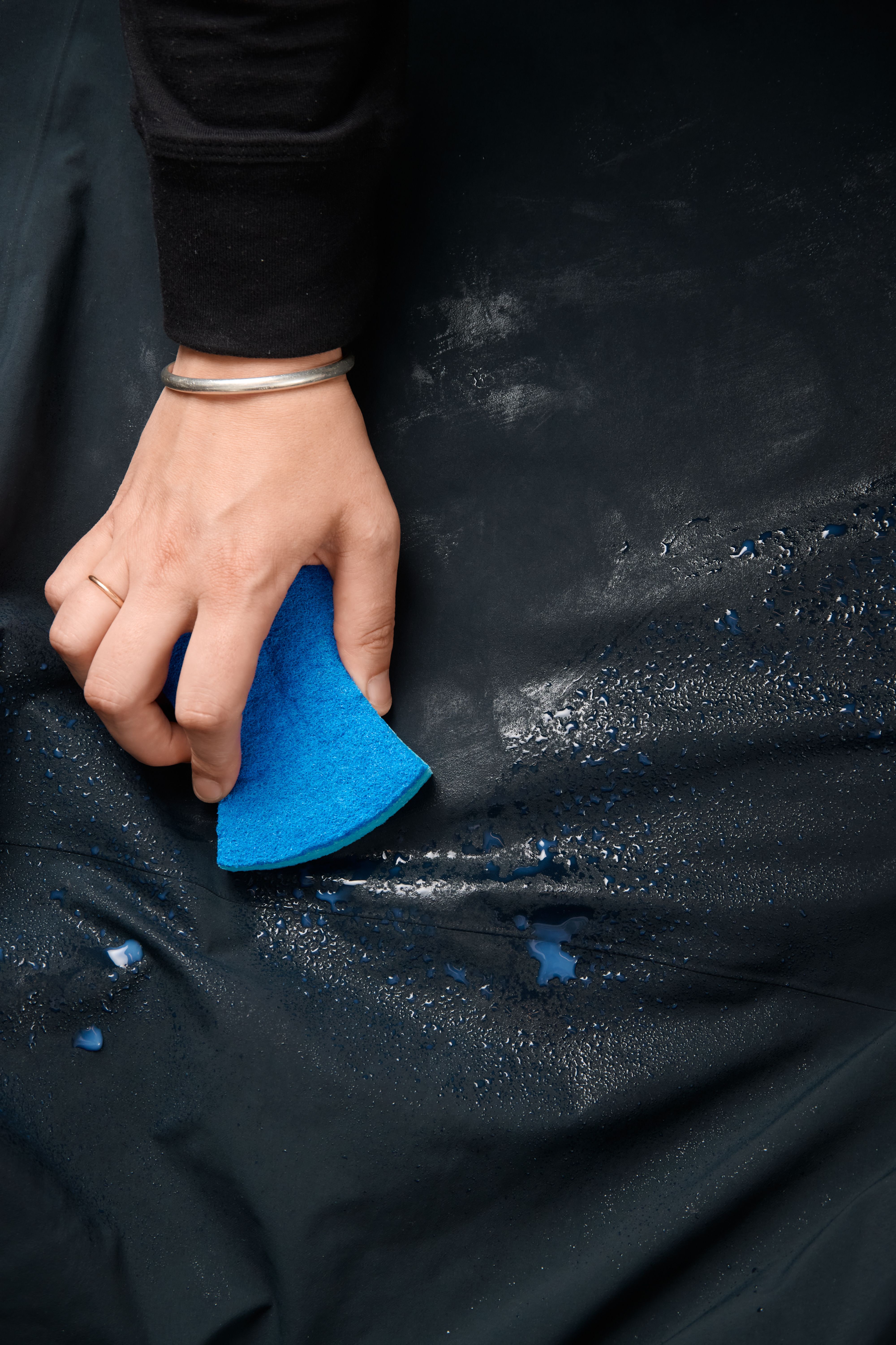 A hand holding a blue sponge wipes water from a dark, possibly waterproof, surface.