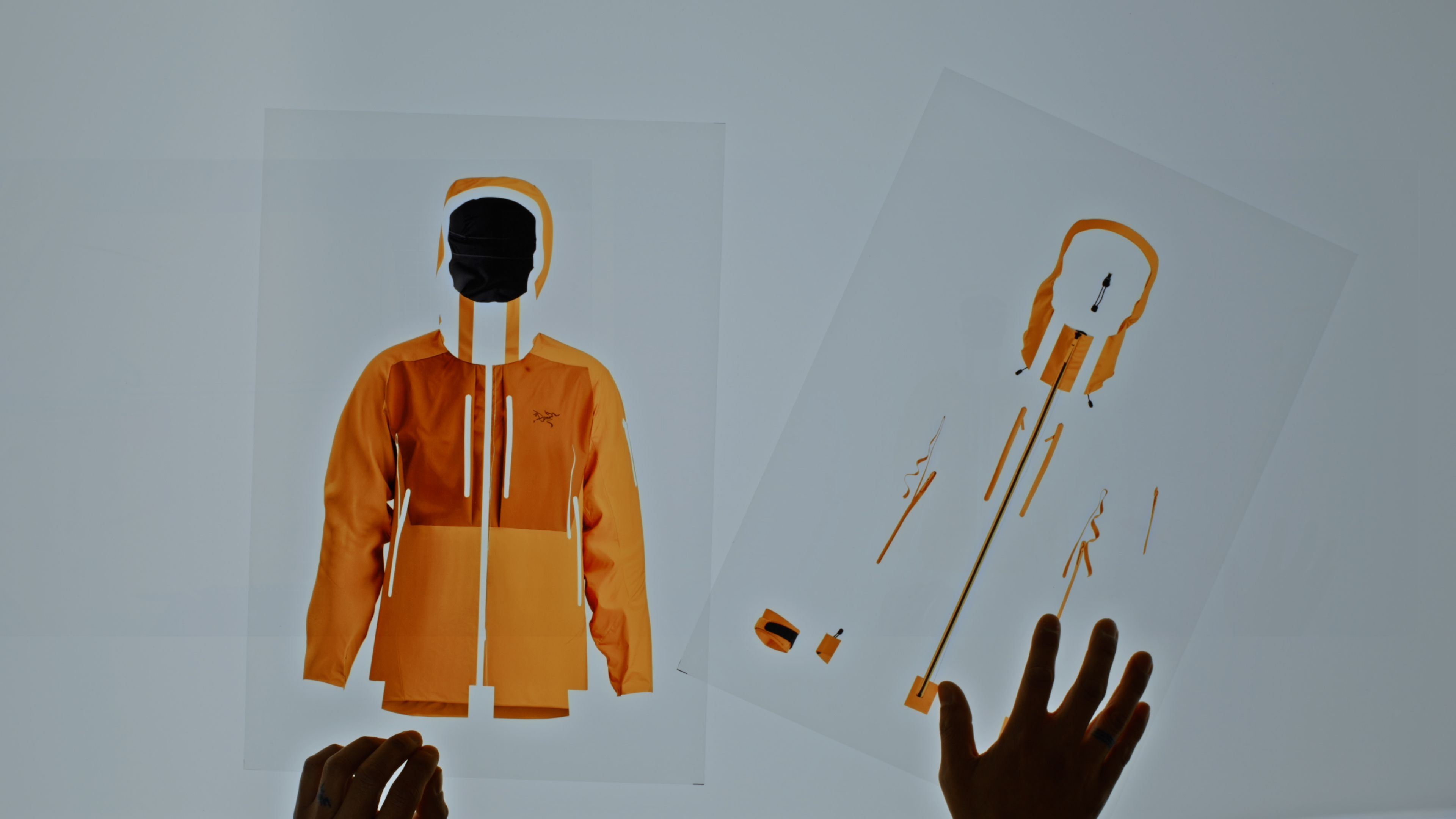 Hands hold two transparent sheets: one showing an orange Arc'teryx jacket, the other its deconstructed parts.