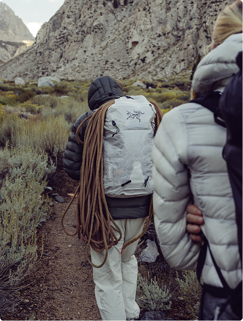 Two hikers from behind walk a rocky mountain trail; one carries a backpack with a coiled climbing rope.