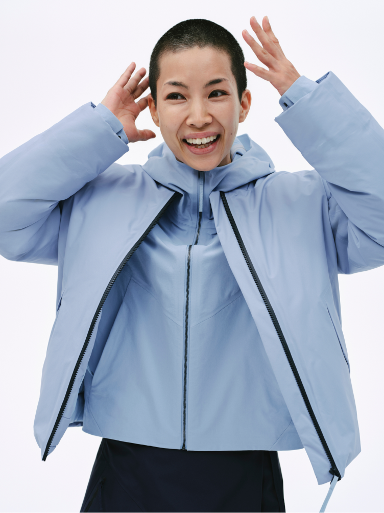 A smiling person with a buzz cut wears two light blue jackets, the outer one partially unzipped, with hands raised by their head.
