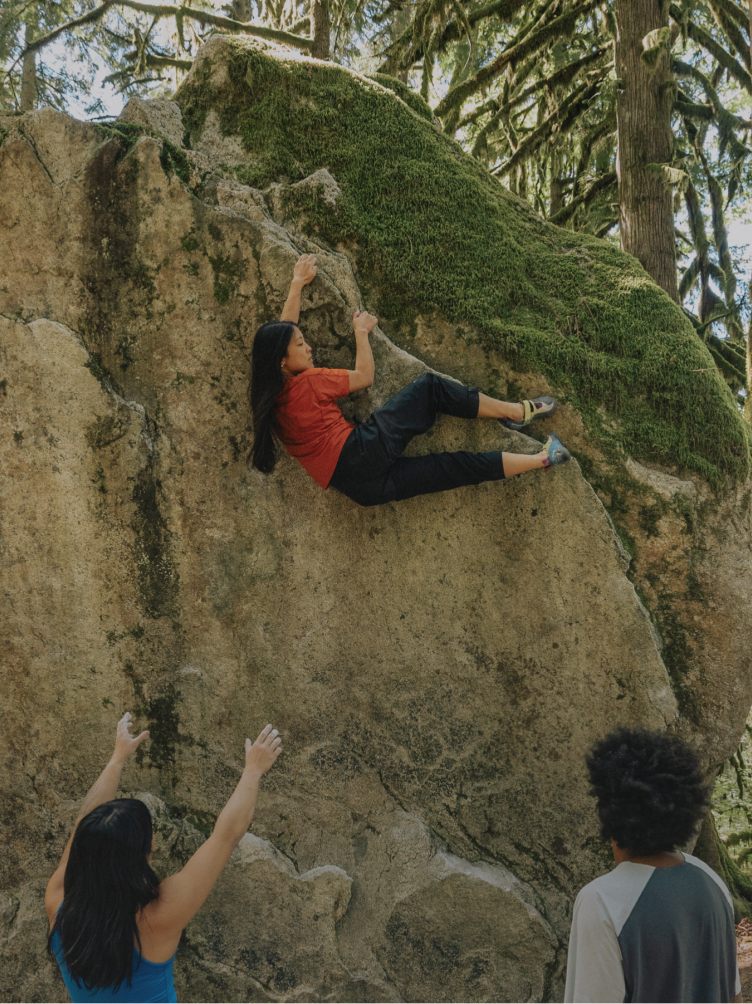 A woman boulders on a mossy rock with two people spotting her below.