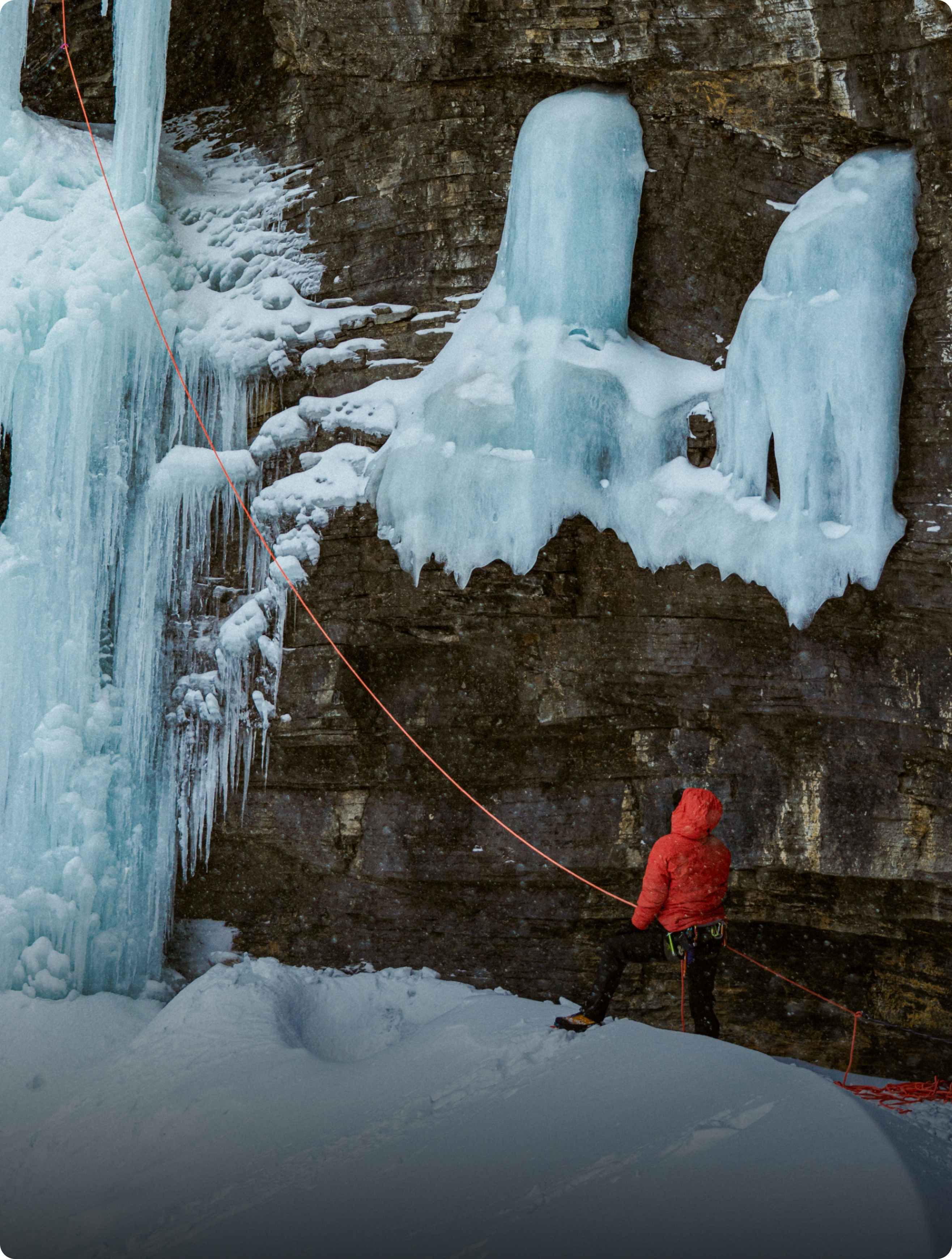 An ice climber in a red jacket stands on snowy ground at the base of a large frozen waterfall.