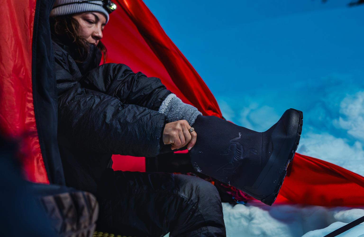 Woman in winter gear puts on a black boot inside a red tent.