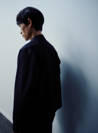 Back view of a person in a dark shirt, head turned left looking down, against a white wall.