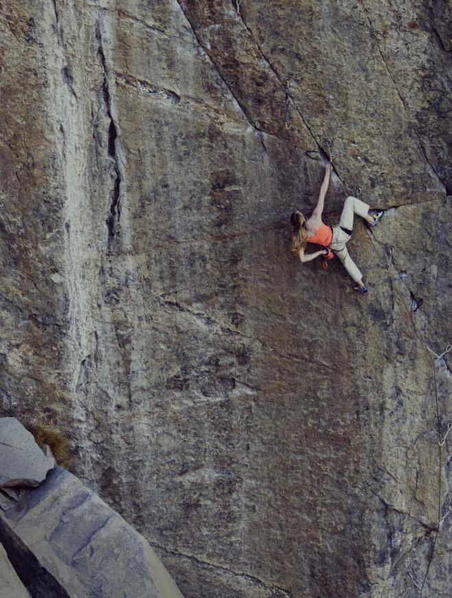 A woman in an orange tank top and light pants rock climbs a textured grey rock face, reaching for a hold.