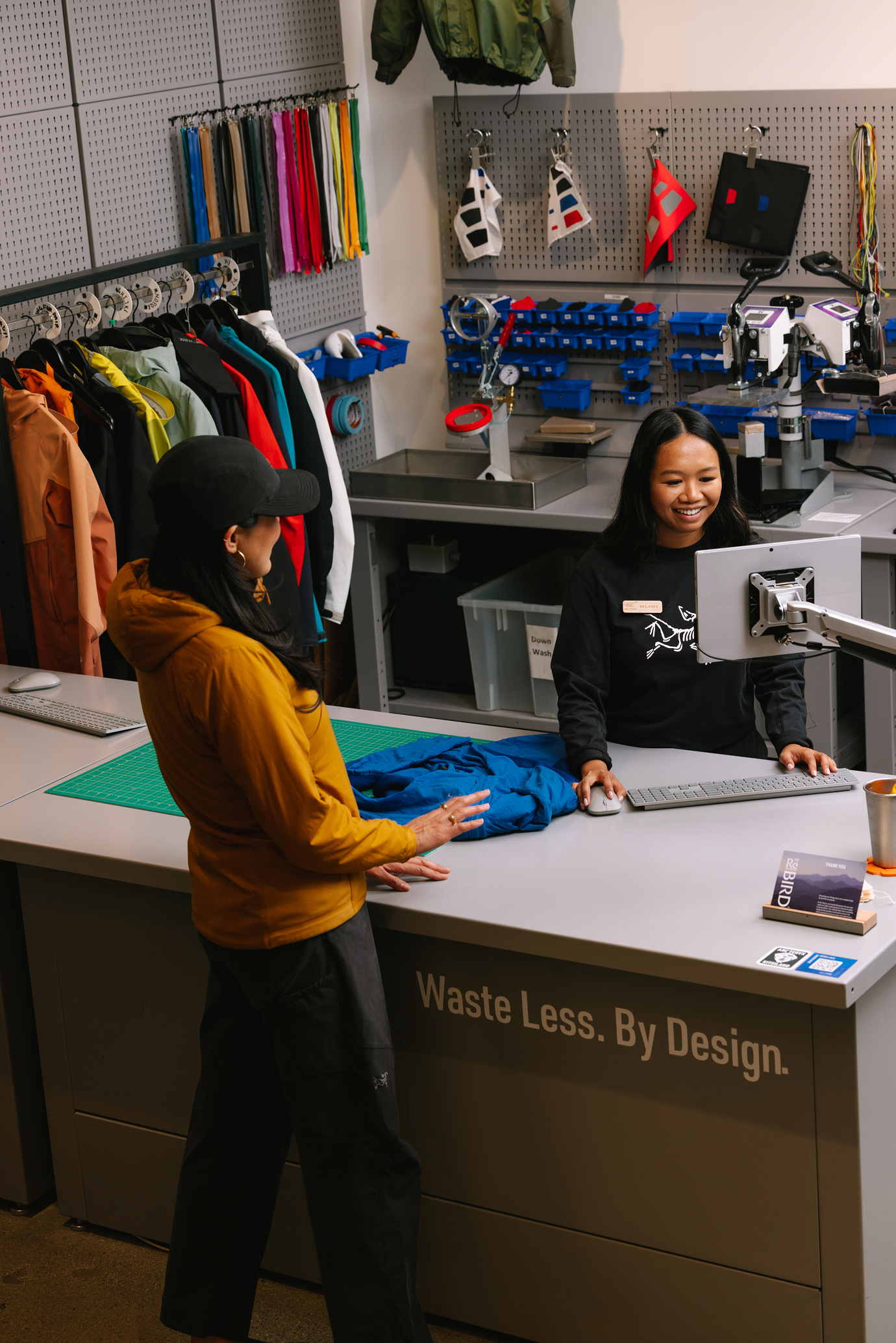 An Arc'teryx employee smiles at a customer over a blue jacket on a counter that says "Waste Less. By Design."