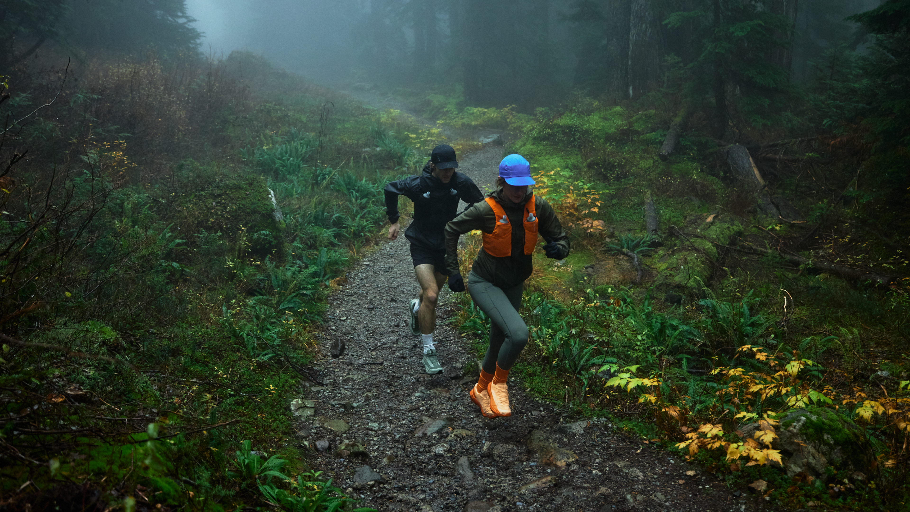 Two people trail running on a muddy path through a misty forest.