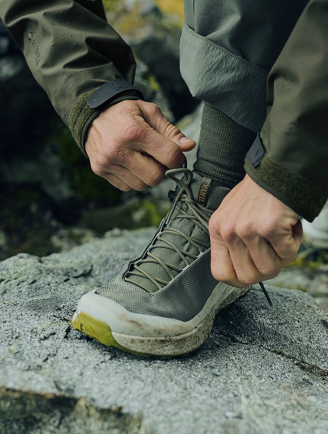 Hands tying the laces of an olive-green Arc'teryx hiking shoe on a rock.