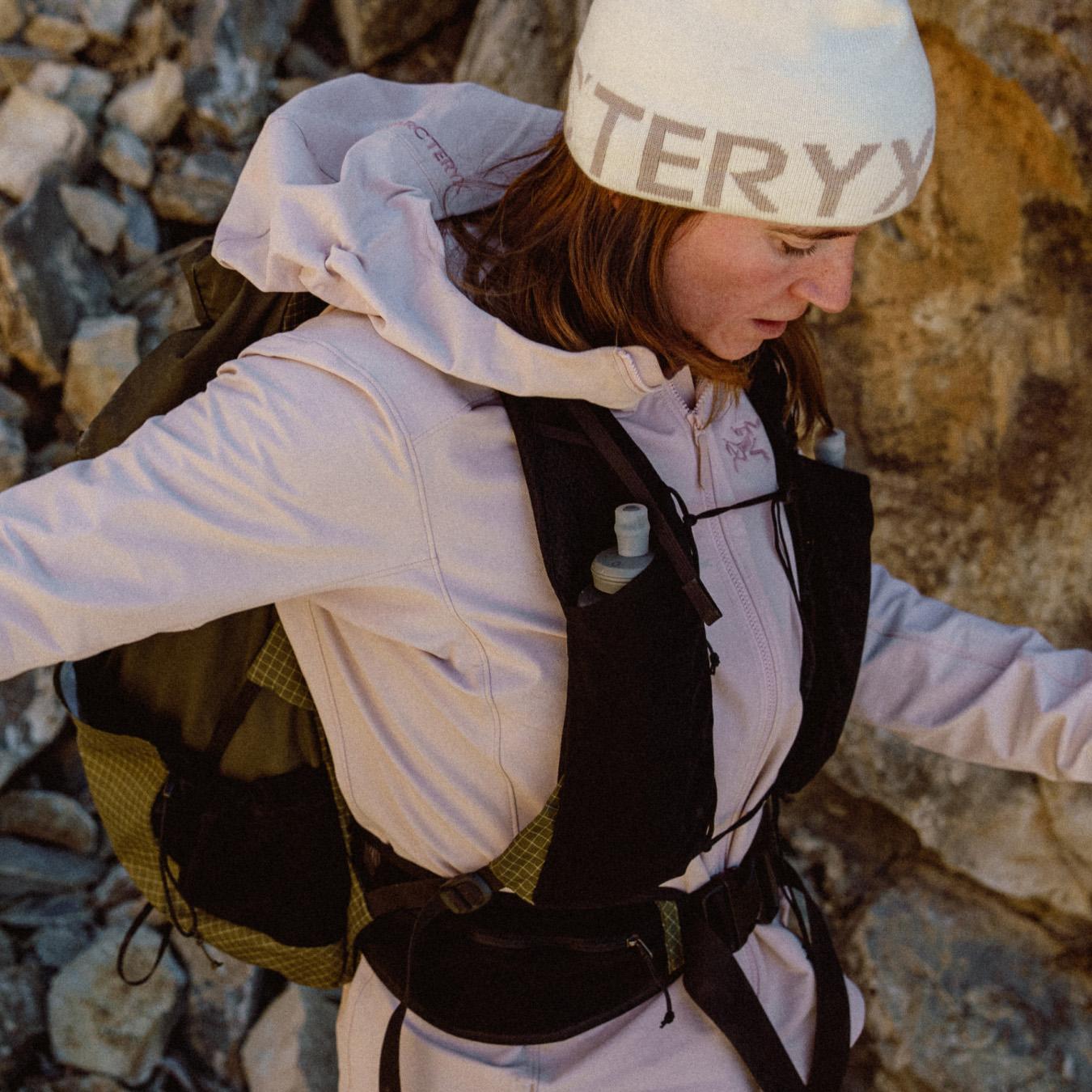 Woman in an Arc'teryx beanie, purple jacket, and hydration vest with a water bottle, in a rocky landscape.