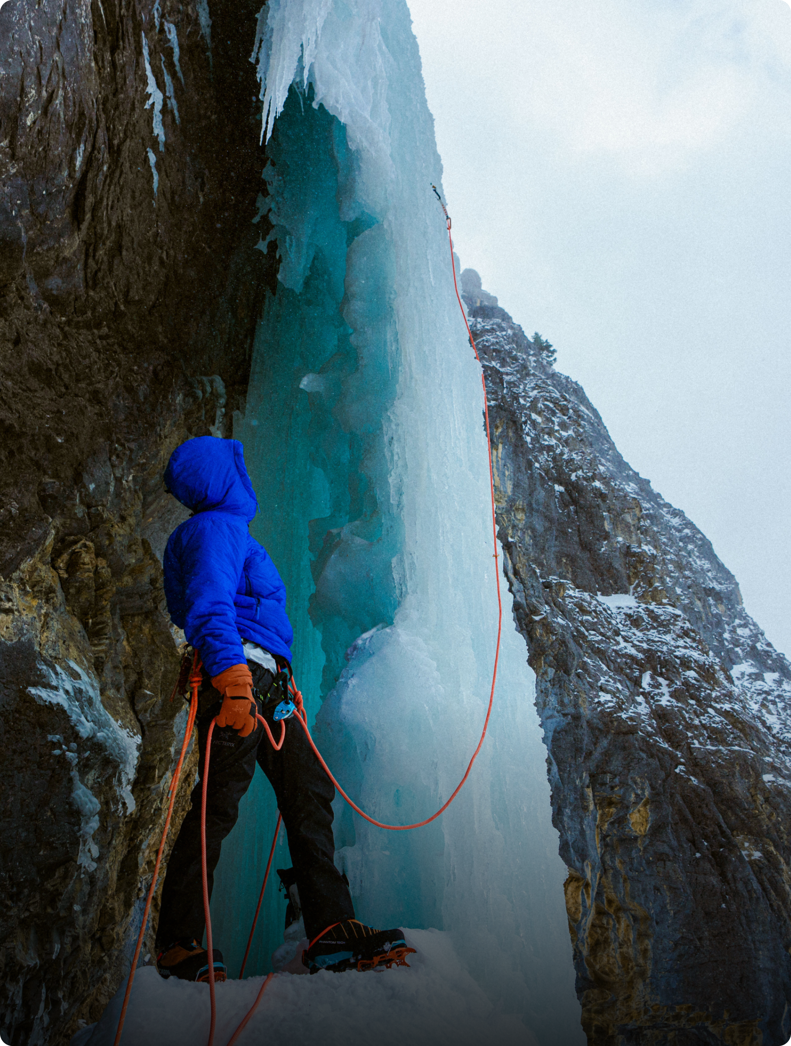 An ice climber in a blue jacket stands at the base of a towering blue ice formation, ready to ascend.