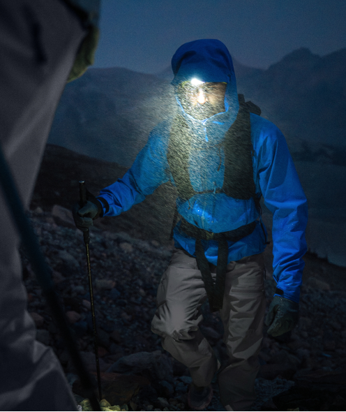 A person in a blue jacket and headlamp hikes at night through falling snow or rain, holding a trekking pole.
