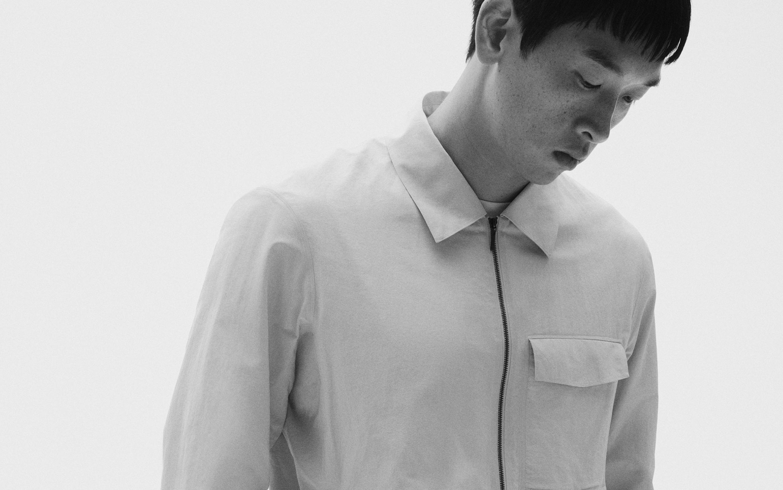 Black and white portrait of a man in a light, collared, zippered jacket, looking down.