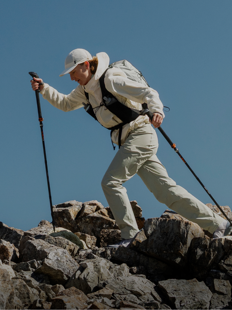 A person in light-colored hiking gear climbs a rocky slope with trekking poles.