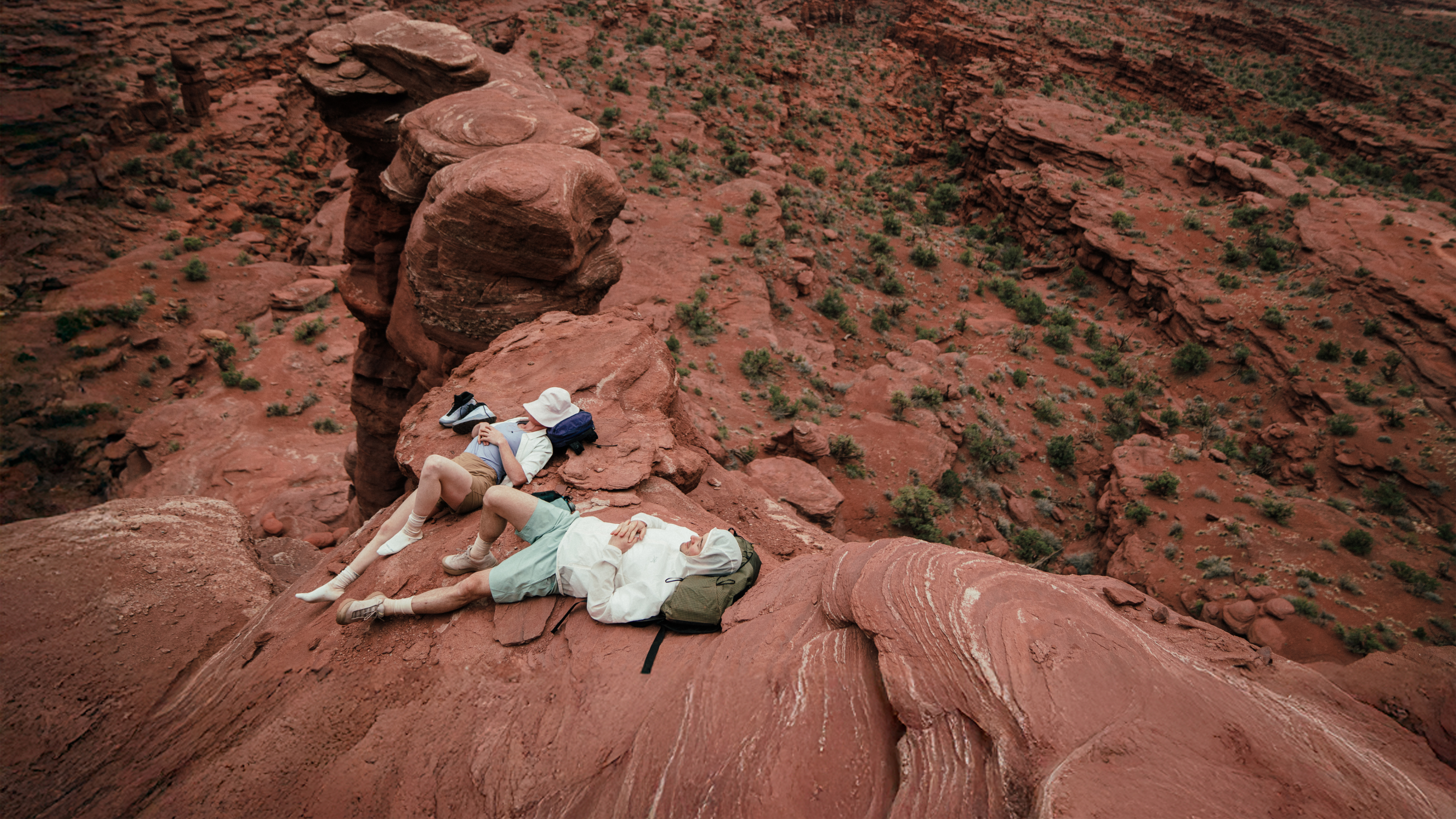 Two people lying on a red rock formation overlooking a vast rocky landscape.