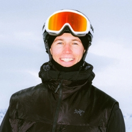 A smiling man wearing a ski helmet, reflective goggles, and a black puffer vest against a snowy background.