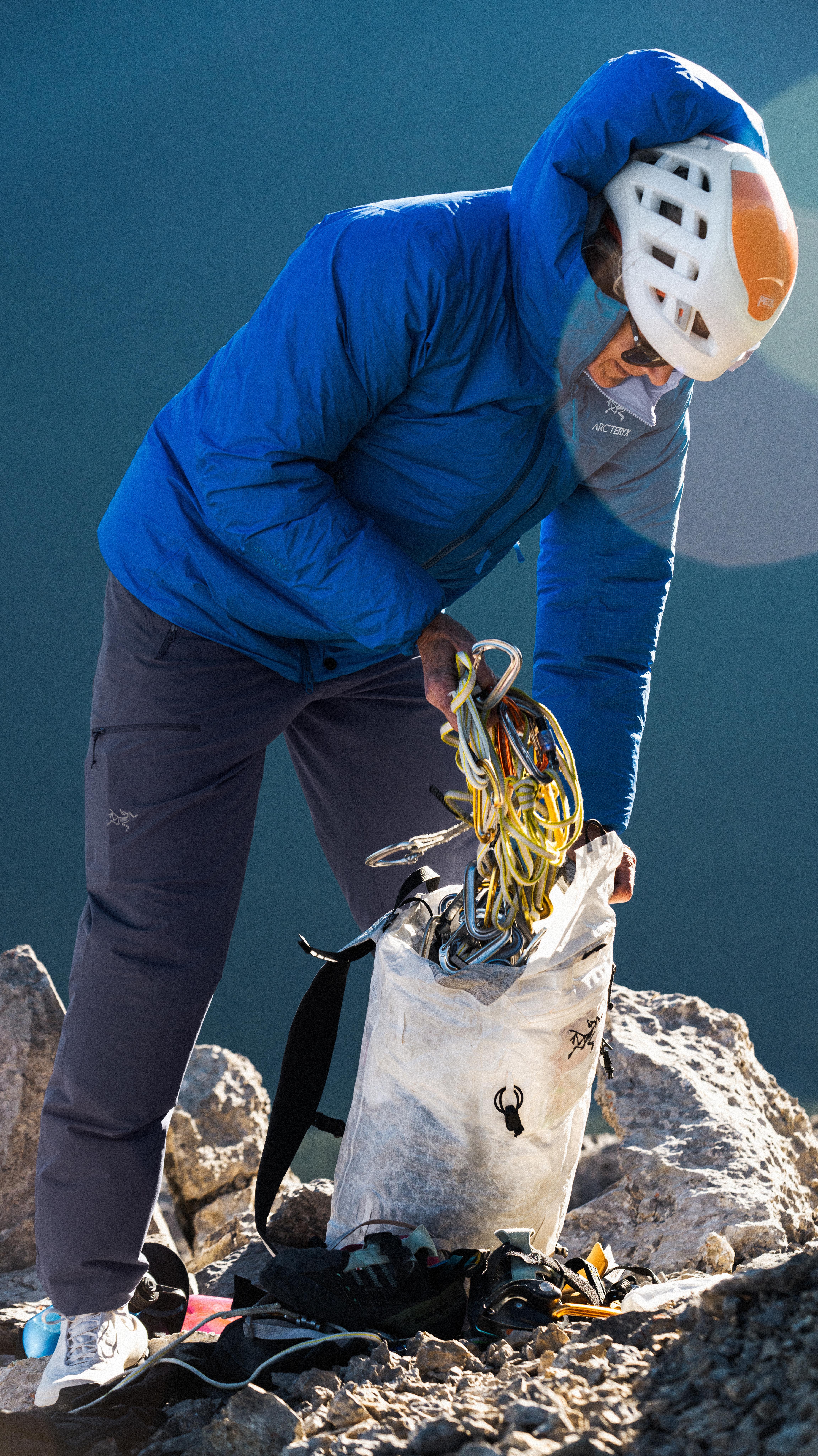 A climber in a blue jacket and helmet puts climbing gear into a white bag on rocky ground.