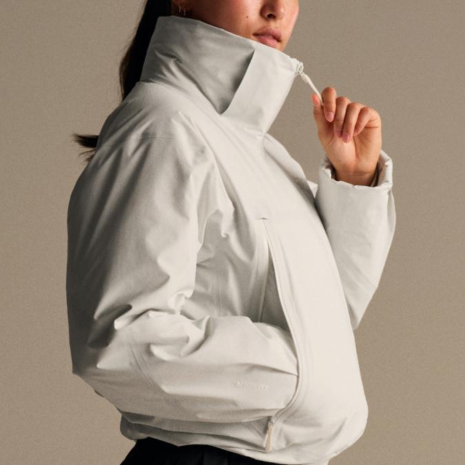 A person wears a light gray technical jacket with a high collar, adjusting the drawstring.