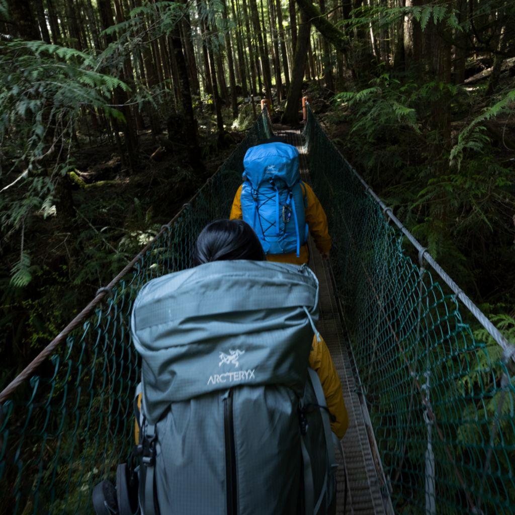 Two hikers with backpacks cross a suspension bridge in a dense forest.
