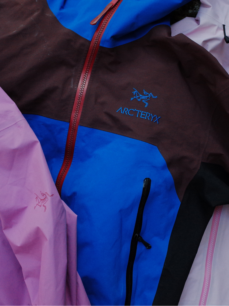 A brown, blue, and black Arc'teryx jacket with a red zipper and blue logo, next to a pink jacket.