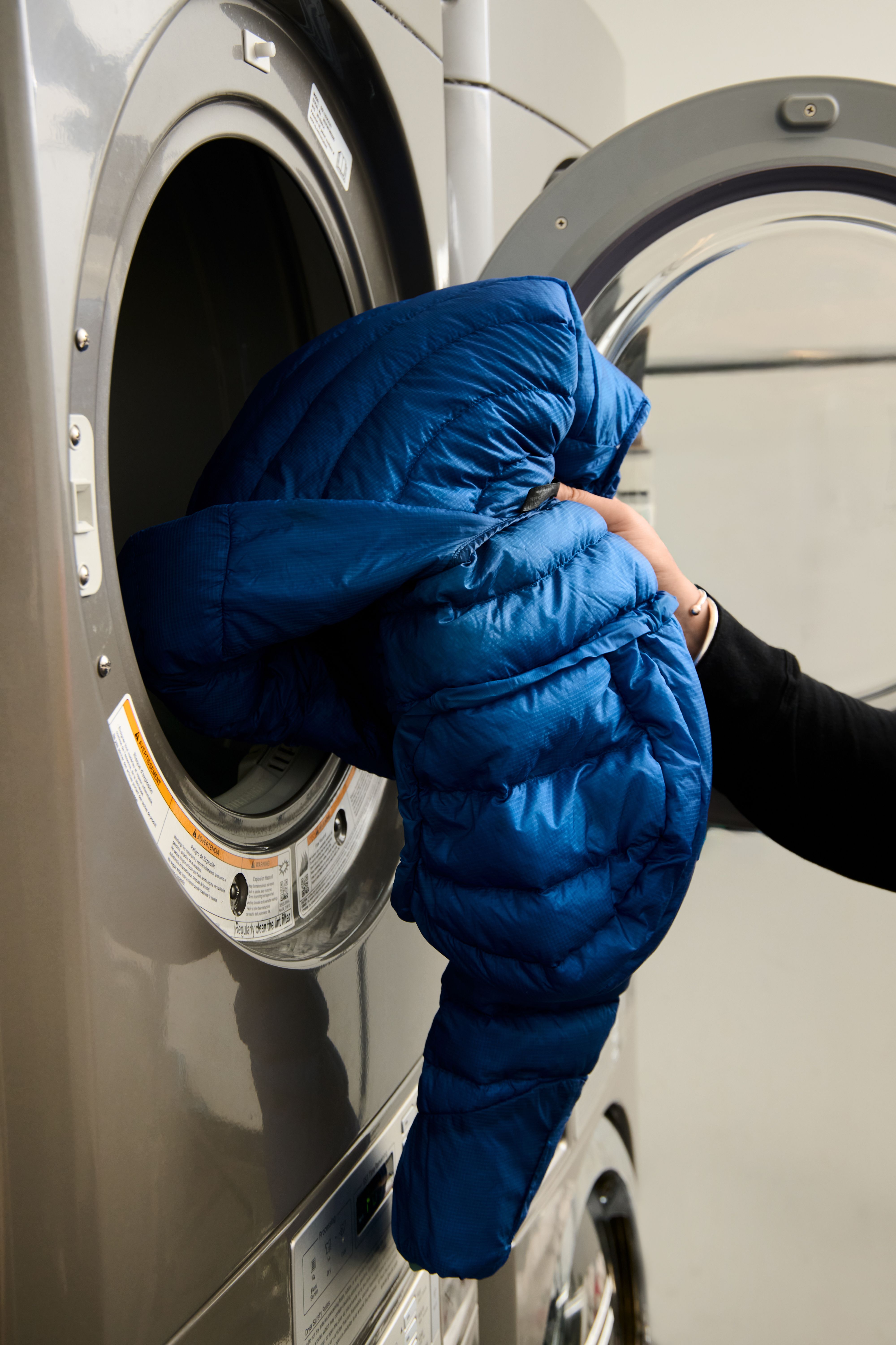 A hand pulls a blue puffer jacket from a front-loading dryer.