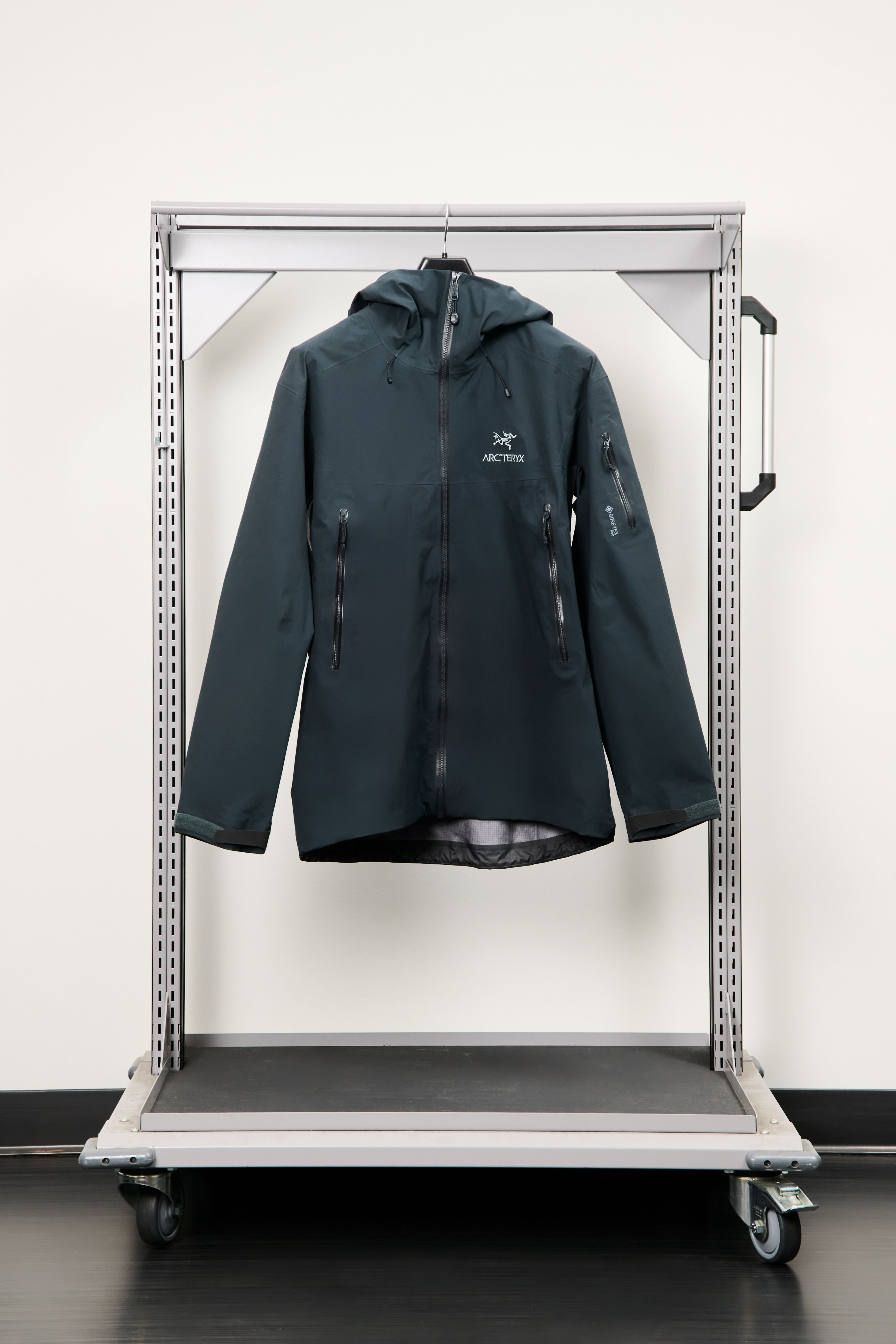 A dark green Arc'teryx hooded jacket hangs on a rolling metal clothing rack.