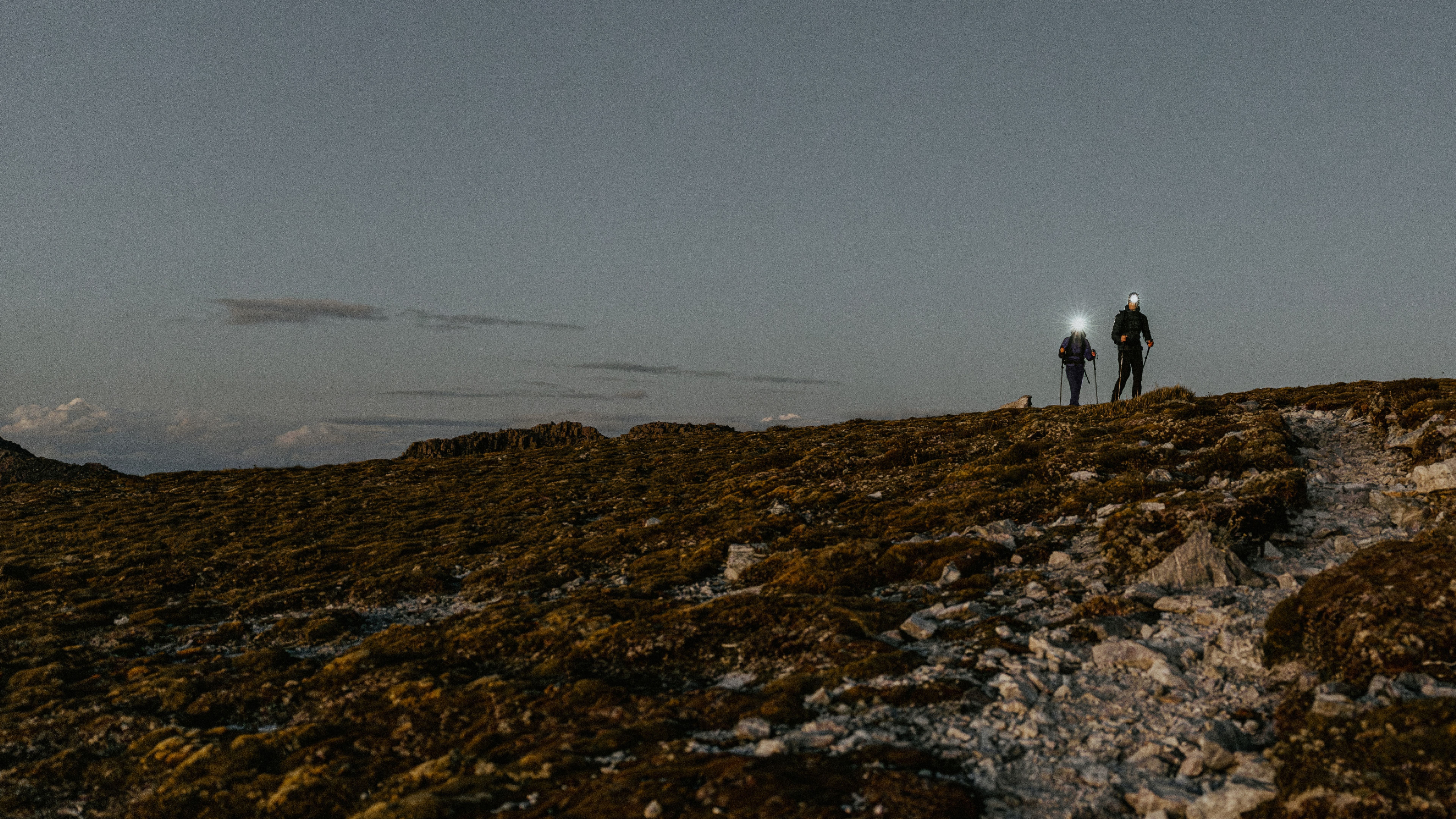 Two hikers with headlamps and trekking poles ascend a rocky, mossy hill at dusk.
