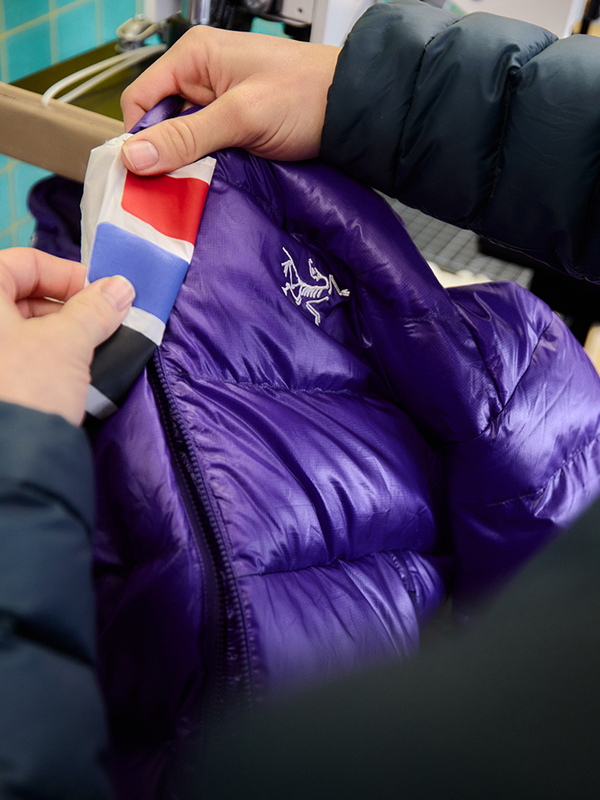 Hands holding a bright purple Arc'teryx puffer jacket with a red, white, and blue striped lining visible at the collar.