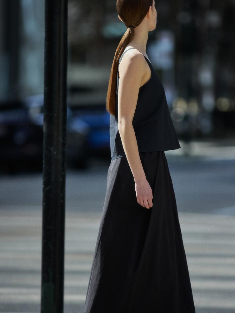 A woman with a long brown ponytail, wearing a black sleeveless top and long black skirt, walks to the right.