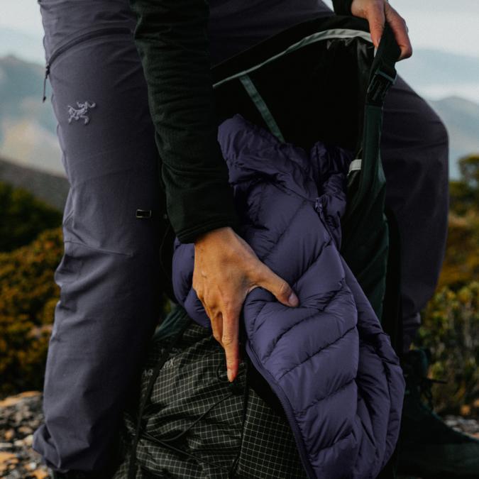 A person puts a purple puffy jacket into a hiking backpack outdoors.