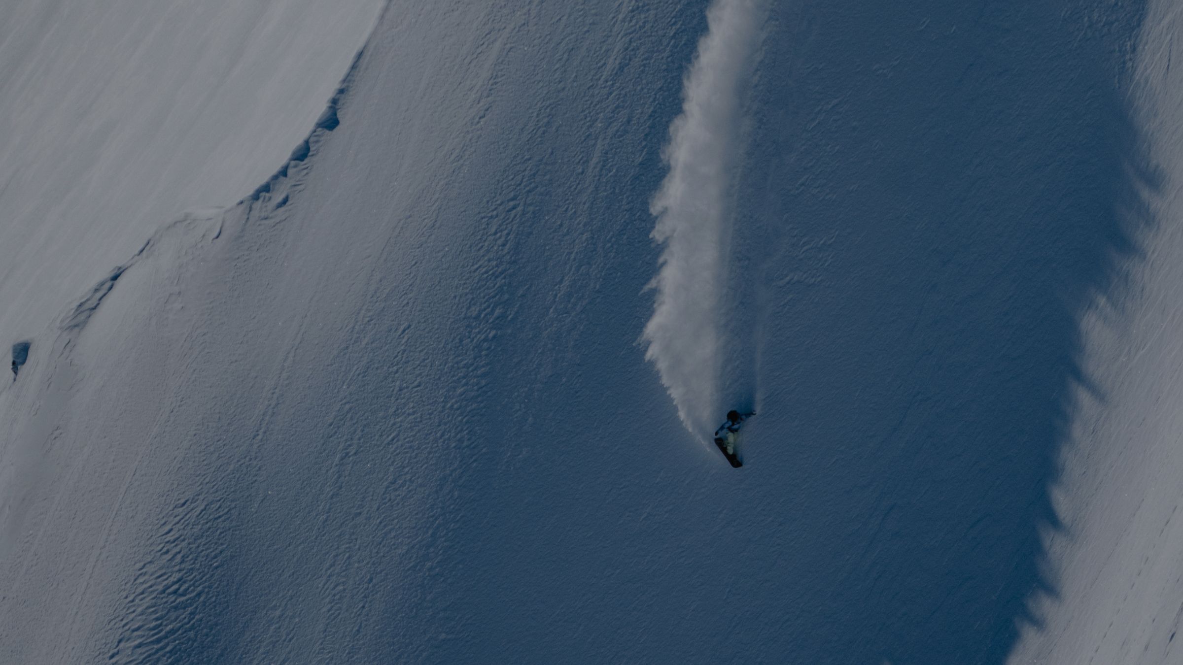 A snowboarder carves down a steep, snow-covered mountain, kicking up a trail of snow.