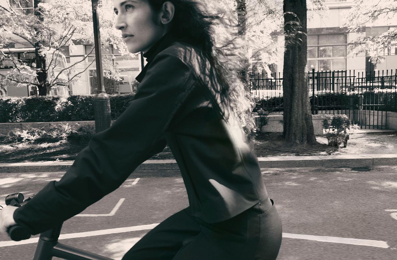 Black and white image of a person with long, flowing hair riding a bicycle on a street.