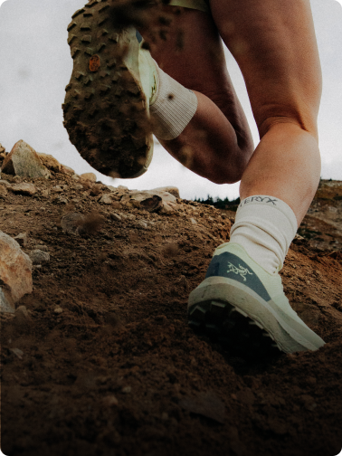 Low-angle view of a runner's legs and trail shoes kicking up dirt on a rocky path.
