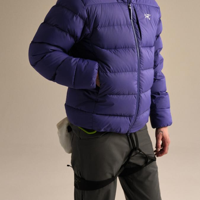 A person wears a purple Arc'teryx puffer jacket, dark pants, and climbing gear.
