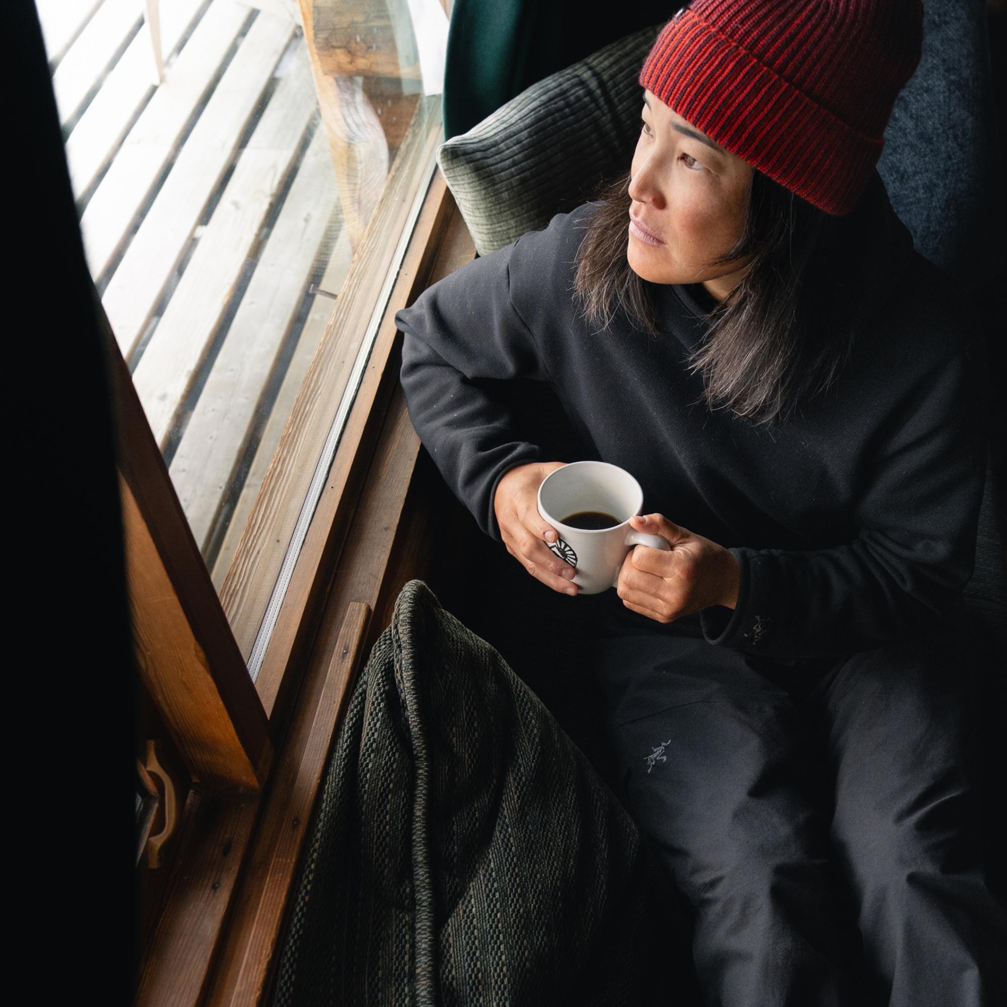A person in a red beanie holds a coffee mug while looking out a window.