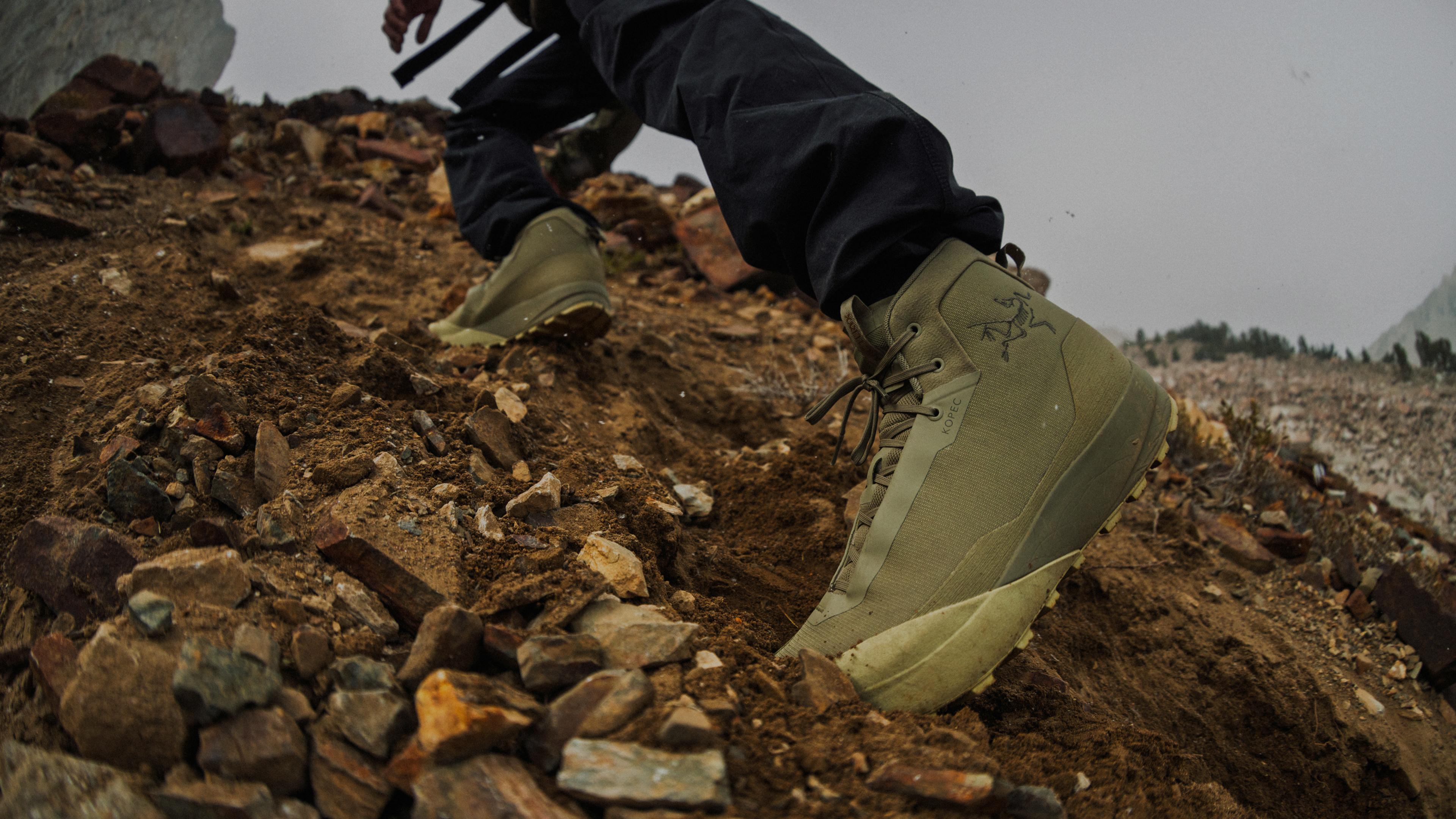 Close-up of a person wearing olive green Arc'teryx hiking boots on a steep, rocky dirt path.