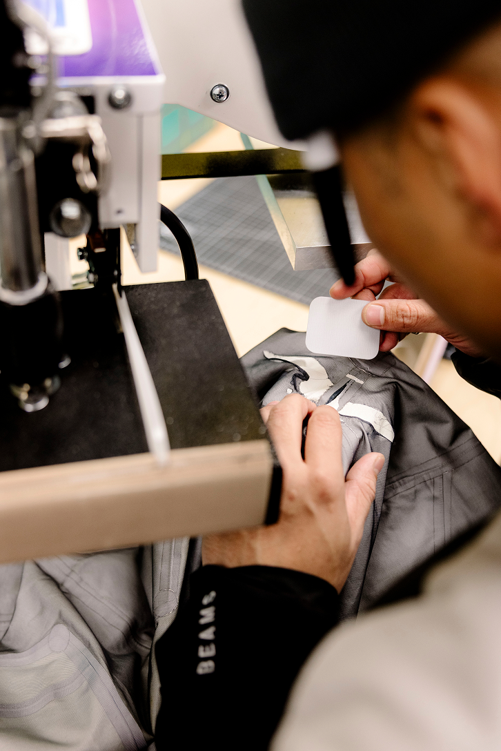 Hands applying a white label to a grey garment with a machine, "BEAMS" visible on the sleeve.