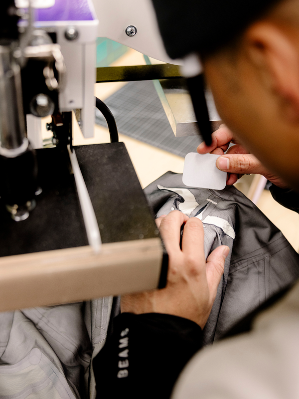 Hands applying a white label to a grey garment with a machine, "BEAMS" visible on the sleeve.