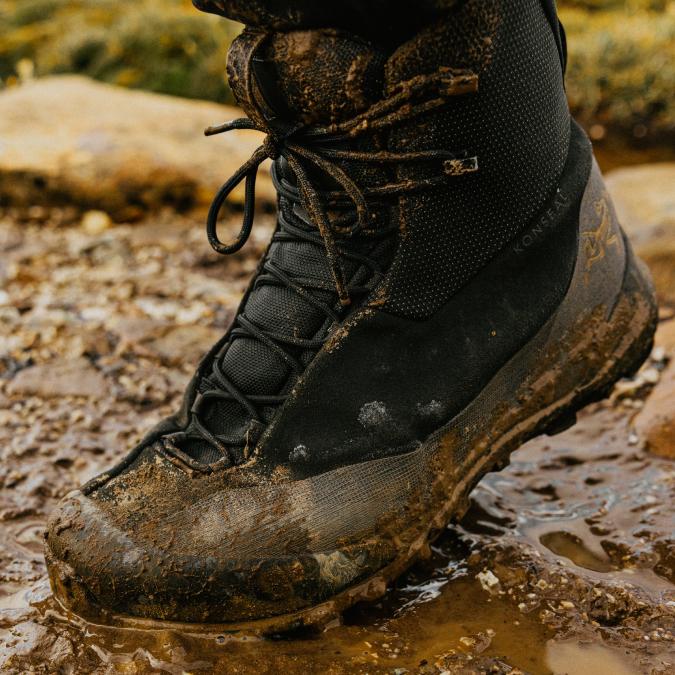 A muddy black Arc'teryx Konseal hiking boot standing in a puddle of mud.