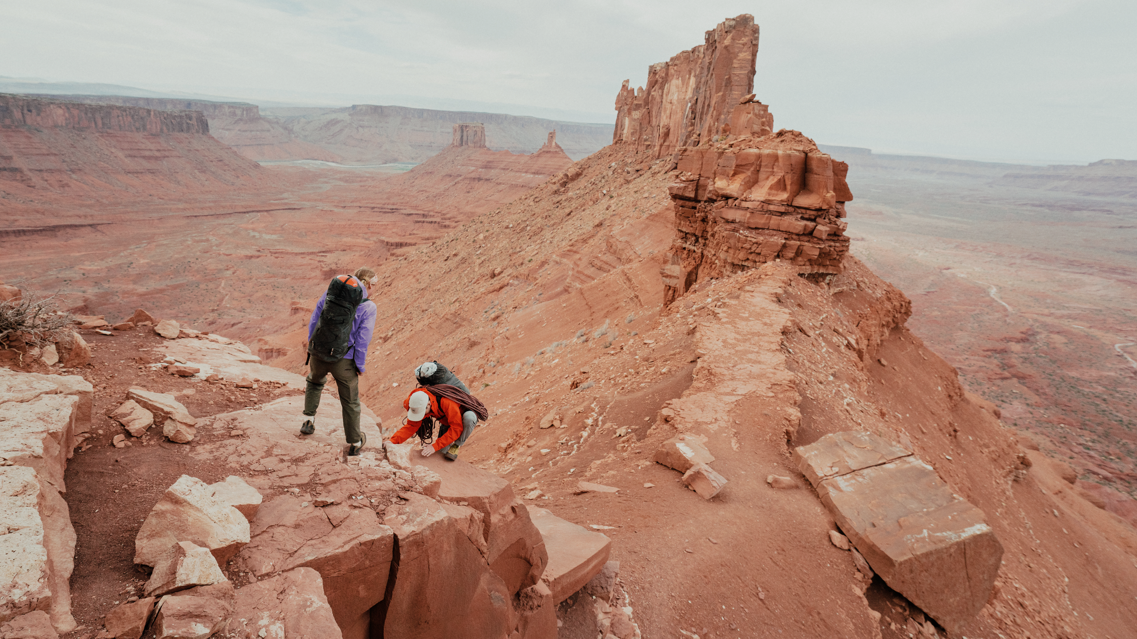 Two hikers on a red rock trail overlooking a vast desert canyon with towering formations.