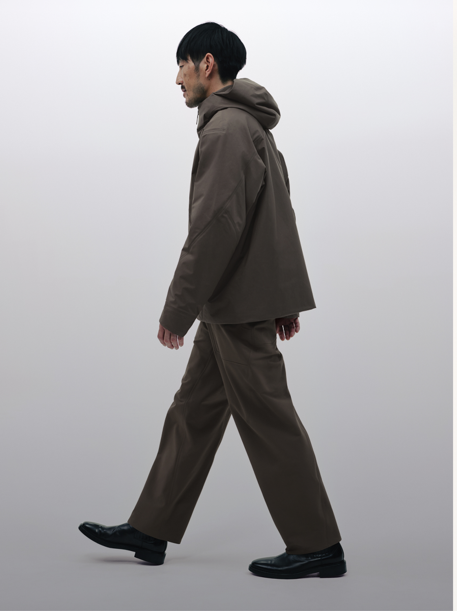 A man walks in side profile, wearing a dark brown hooded jacket, wide-leg trousers, and black shoes against a light gray background.