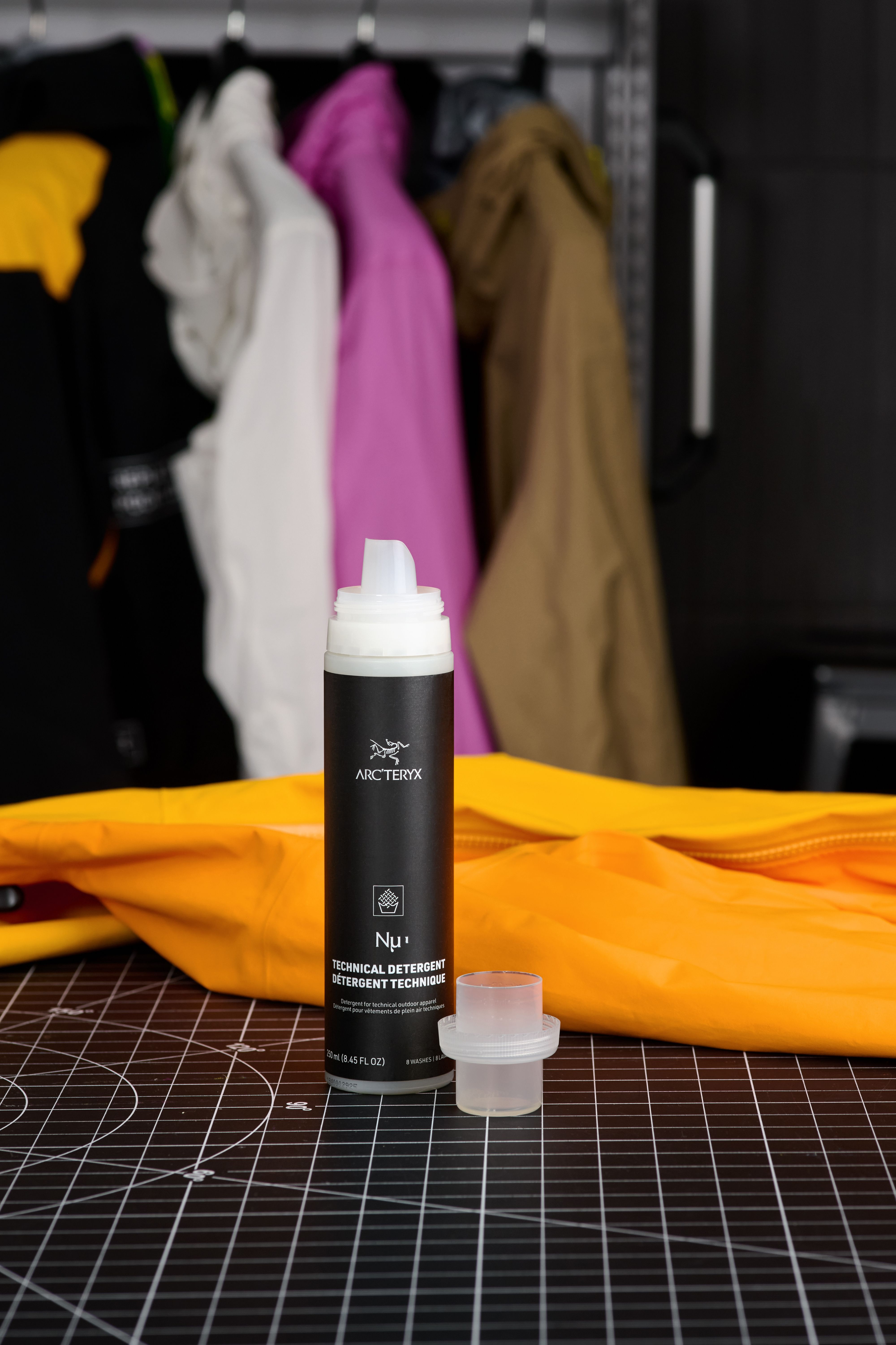 An Arc'teryx Nu technical detergent bottle and measuring cup on a black cutting mat, with an orange jacket and blurred hanging jackets in the background.