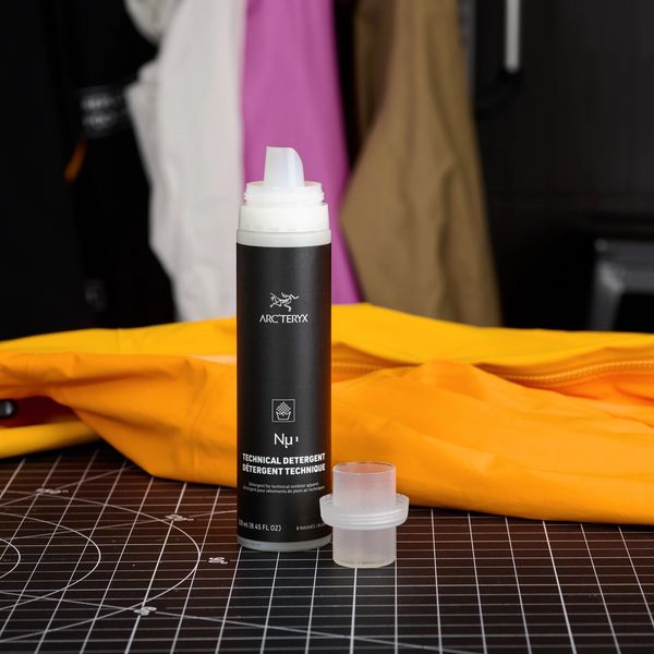 An Arc'teryx Nu technical detergent bottle and measuring cup on a black cutting mat, with an orange jacket and blurred hanging jackets in the background.