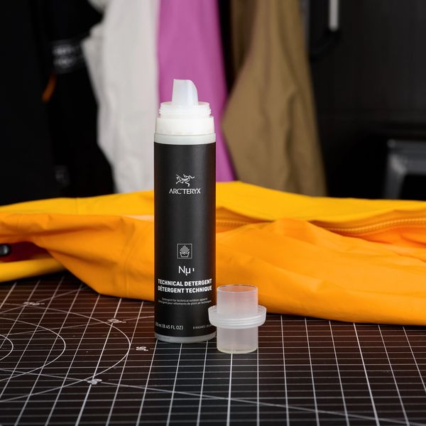 Arc'teryx Nu'1 Technical Detergent bottle and measuring cup beside a yellow jacket on a black grid mat, with blurred hanging jackets in the background.
