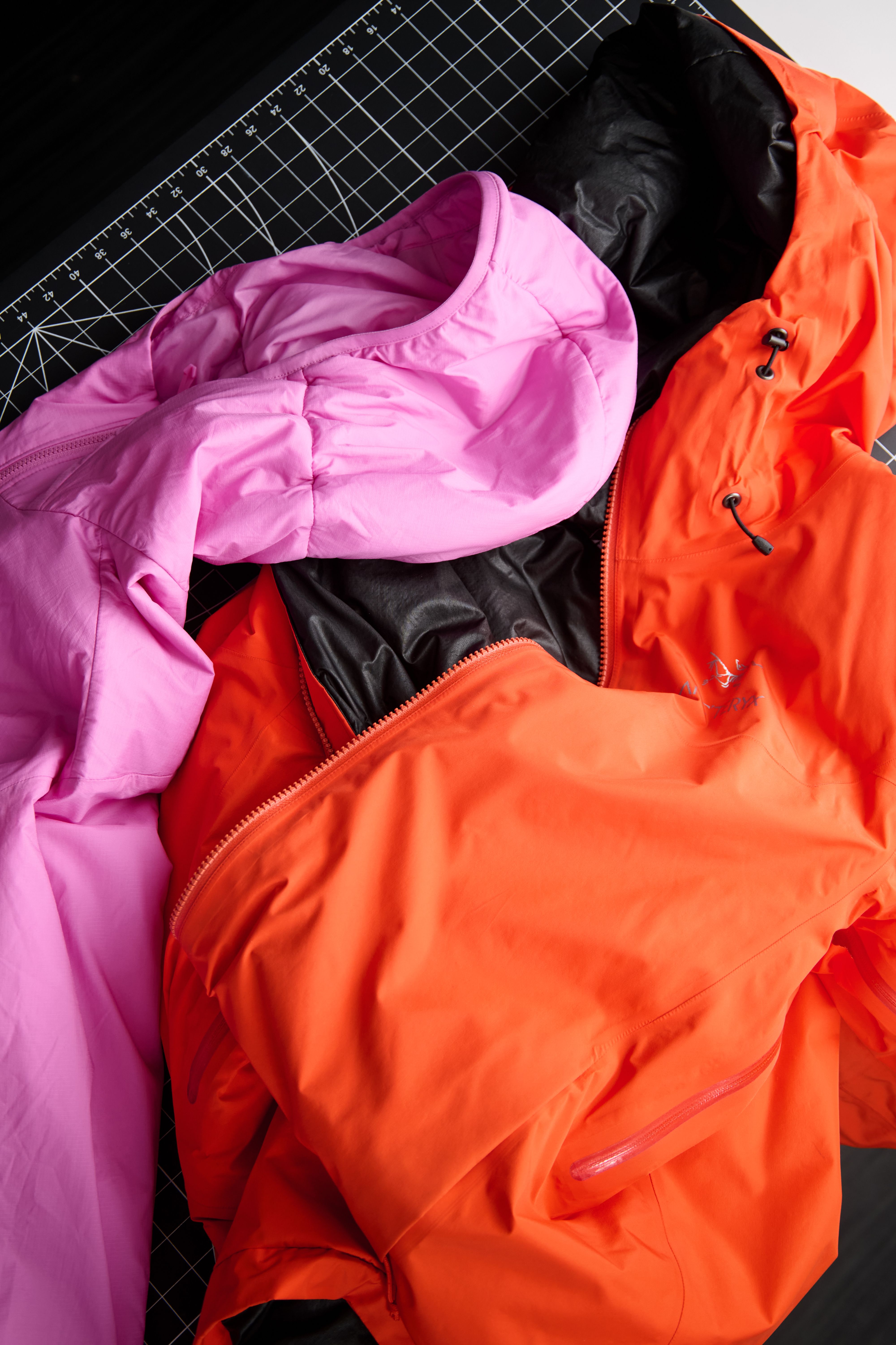 Pink, orange, and black jackets laid on a black grid mat.