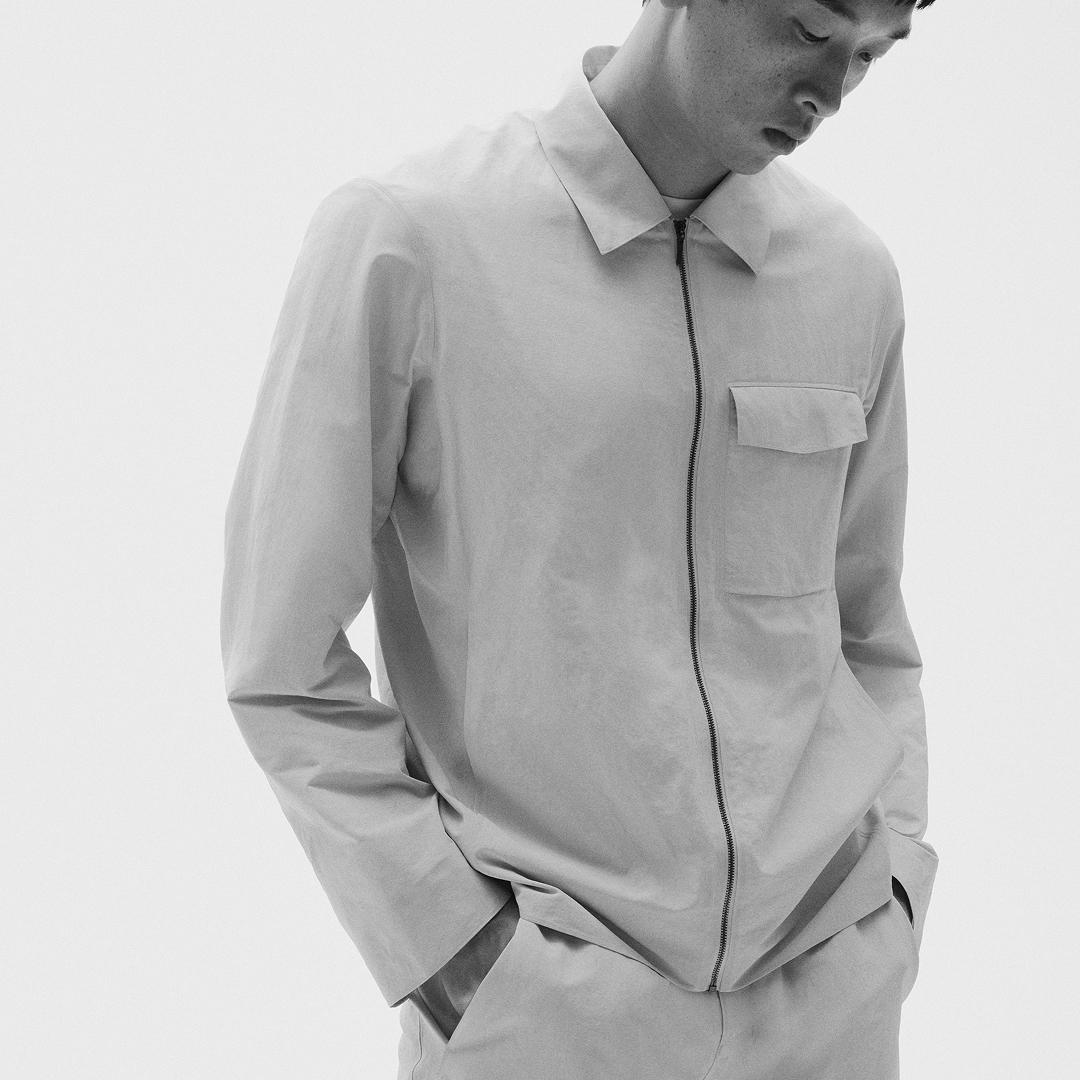 Monochromatic image of a man wearing a light collared jacket with a zipper and chest pocket, hands in pockets, looking down.